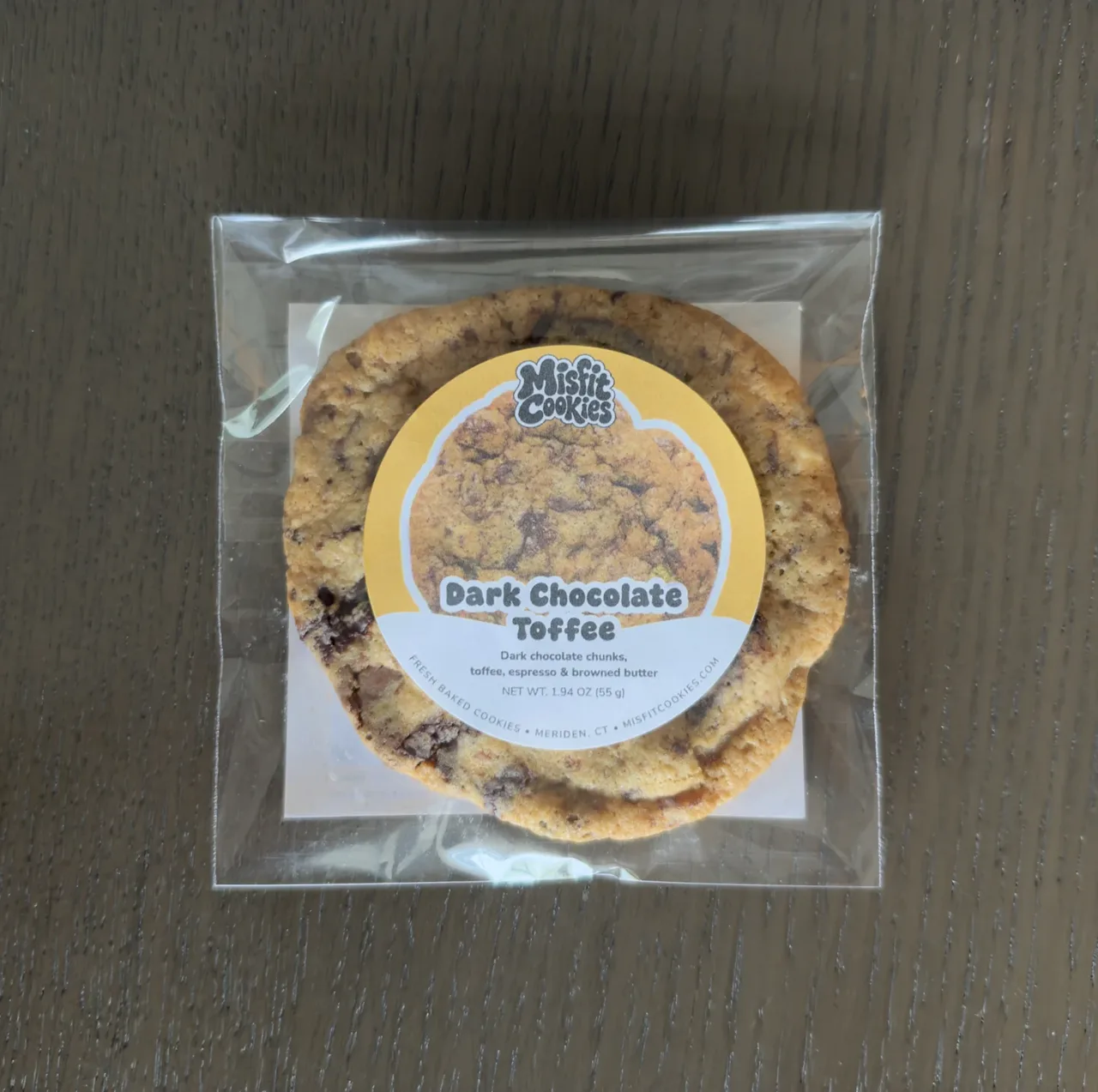 A packaged Dark Chocolate Toffee cookie with its golden yellow Misfit Cookies label.